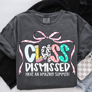 May include: A dark gray t-shirt with the text "Class Dismissed Have an Amazing Summer!" in pink, white, and black lettering. The word "Class" is decorated with colorful patterns and a pink bow.