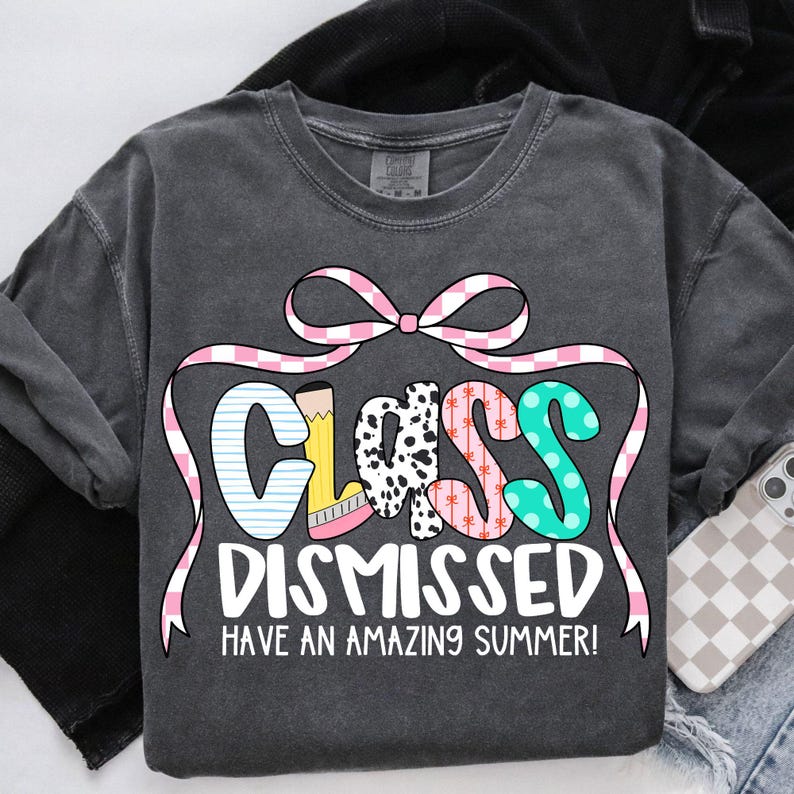 May include: A dark gray t-shirt with the text "Class Dismissed Have an Amazing Summer!" in pink, white, and black lettering. The word "Class" is decorated with colorful patterns and a pink bow.