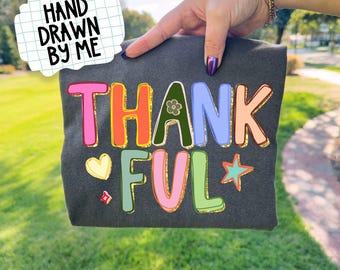 May include: Dark gray t-shirt with the word "THANKFUL" in colorful, outlined letters. A small heart, flower, and star accent the design. The phrase "HAND DRAWN BY ME" is in a speech bubble.
