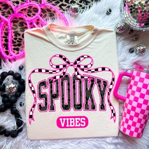 May include: Cream-colored t-shirt with the words "SPOOKY VIBES" in black and pink lettering. The design includes a checkered bow and ribbon. A pink checkered tumbler is visible in the background.