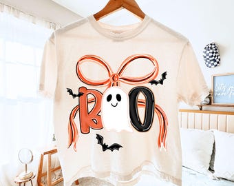May include: A cream-colored t-shirt with a Halloween-themed design. The word "BOO" is displayed in orange and black, with a white ghost in the center. Black bats are scattered around the design. The shirt is hanging on a wooden hanger.