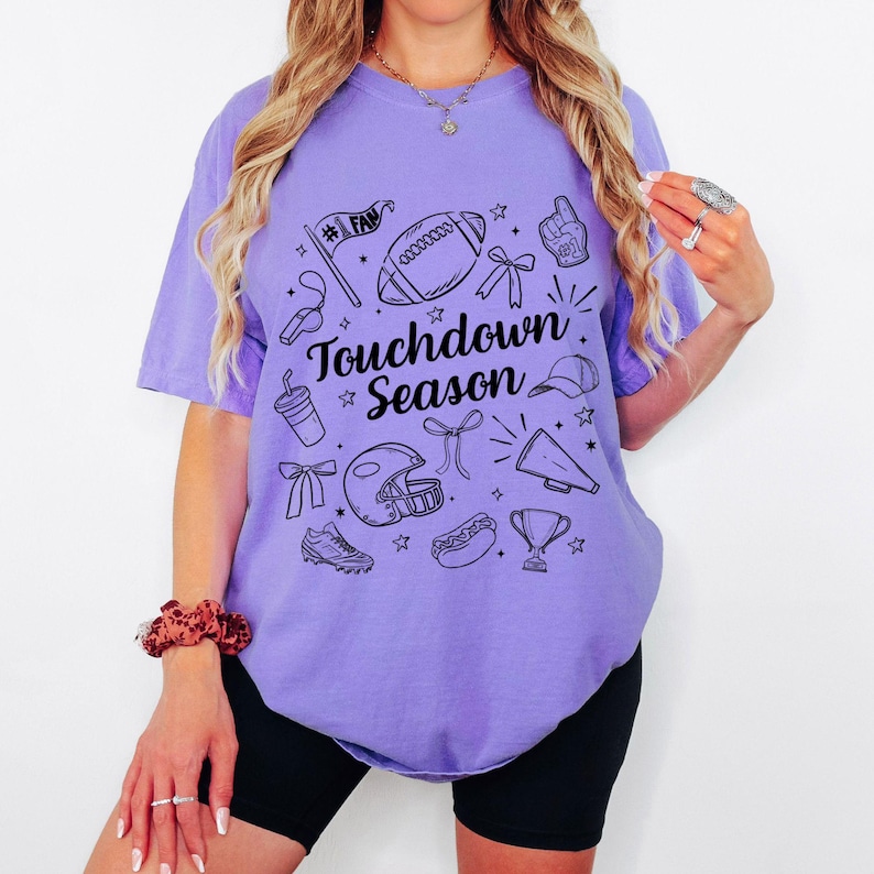 May include: A lilac-colored t-shirt with a black graphic design featuring football-related icons and the text "Touchdown Season." The design includes a football, helmet, trophy, and other sports-themed elements.