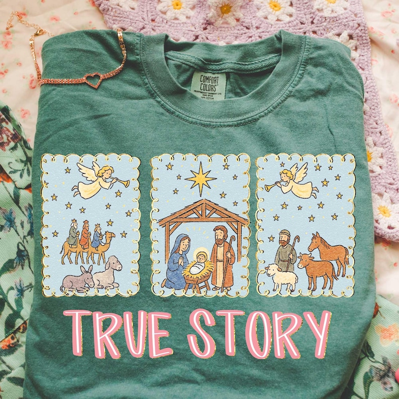 May include: A teal Comfort Colors t-shirt featuring a nativity scene with angels, stars, and the text "TRUE STORY" in pink. The design includes illustrations of the three wise men, the holy family, and shepherds.