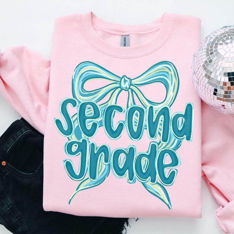 Op de afbeelding: Een lichtroze sweatshirt met de tekst "Second Grade" in turquoise script. Een grote turquoise en lichtblauwe strik-afbeelding staat boven de tekst. Een zilveren discobal staat op de achtergrond.