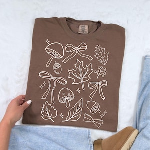 Op de afbeelding: Een bruin T-shirt met een witte afbeelding van herfstthema's, waaronder paddenstoelen, strikken, bladeren en eikels. Het shirt is van het merk Comfort Colors, maat XL, en is gemaakt van een zacht materiaal.