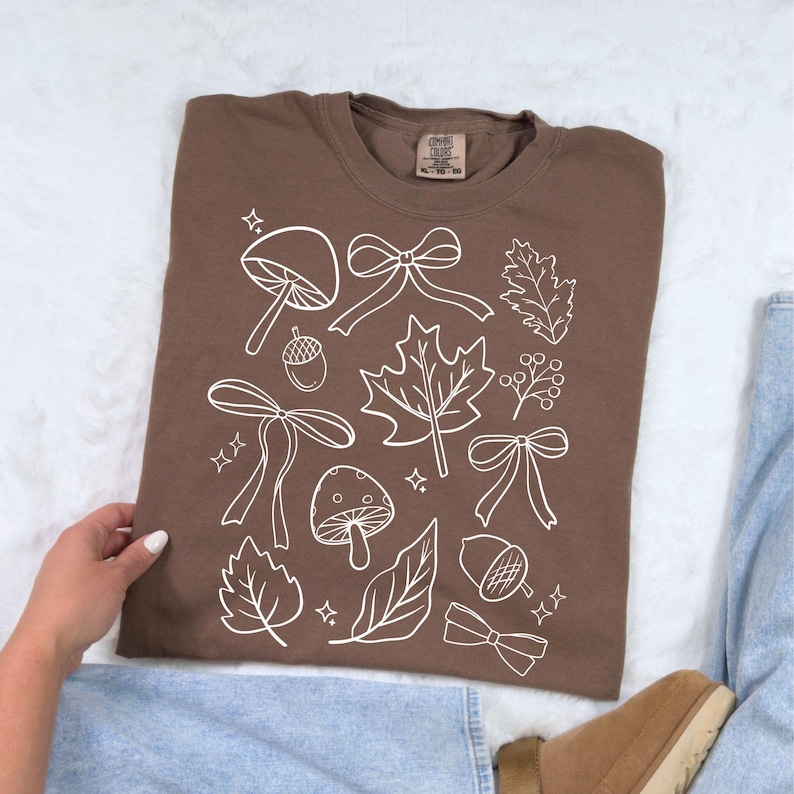 Op de afbeelding: Een bruin T-shirt met een witte afbeelding van herfstthema's, waaronder paddenstoelen, strikken, bladeren en eikels. Het shirt is van het merk Comfort Colors, maat XL, en is gemaakt van een zacht materiaal.