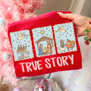 May include: Red t-shirt featuring a nativity scene design with the text "TRUE STORY". The design includes illustrations of angels, the three wise men, and the holy family. The illustrations are set against a light blue background.