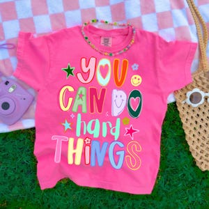 May include: A bright pink t-shirt with the phrase "YOU CAN DO HARD THINGS" in colorful, playful lettering. The shirt features stars, flowers, and a smiley face graphic. A beaded necklace and a woven bag are also visible.