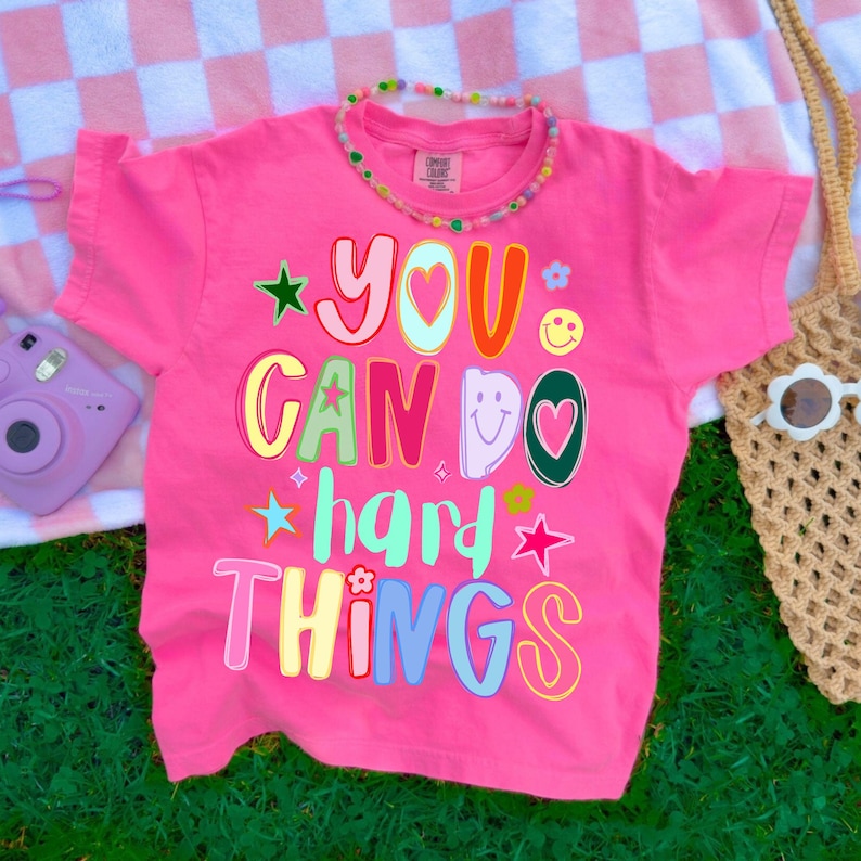 May include: A bright pink t-shirt with the phrase "YOU CAN DO HARD THINGS" in colorful, playful lettering. The shirt features stars, flowers, and a smiley face graphic. A beaded necklace and a woven bag are also visible.