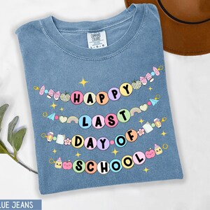 May include: A blue jean t-shirt with the text "Happy Last Day of School" in a colorful banner design. The banner is made of circles with different school-related icons and text.