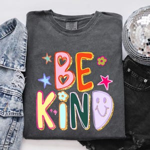 May include: Dark gray sweatshirt with the words "BE KIND" in colorful, retro-style lettering. The design includes stars, flowers, and a smiley face. The sweatshirt is surrounded by a denim jacket, a disco ball, and black shorts.