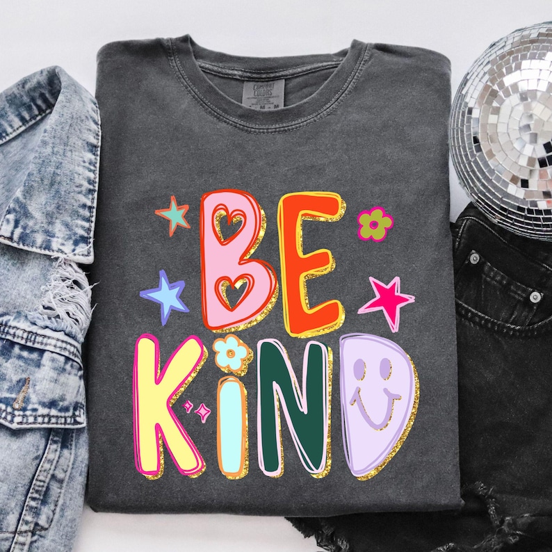 May include: Dark gray sweatshirt with the words "BE KIND" in colorful, retro-style lettering. The design includes stars, flowers, and a smiley face. The sweatshirt is surrounded by a denim jacket, a disco ball, and black shorts.