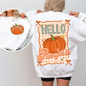 Może przedstawiać: Biała bluza z nadrukiem dyni. Z przodu mała dynia z kokardą. Z tyłu większy wzór dyni z napisem "HELLO Pumpkin" w jesiennej czcionce.