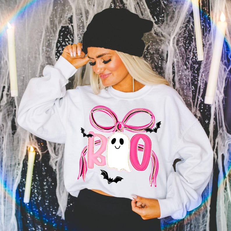 May include: White sweatshirt with a pink bow and the word "BOO" in pink letters, with a ghost and bats. The person in the image is wearing a black beanie.