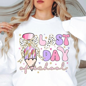 Op de afbeelding: Wit sweatshirt met een roze, paarse en gouden potlood met een strik erop. De tekst "Laatste schooldag" staat in roze, paarse en witte letters.
