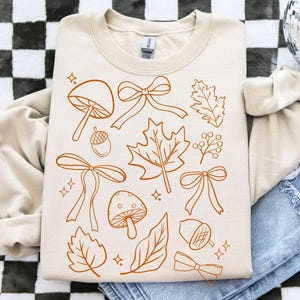 Op de afbeelding: Cr&egrave;mekleurige sweatshirt met een herhalend patroon van herfstthema-illustraties in een warme bruine tint. Het ontwerp bevat paddenstoelen, strikken, bladeren, eikels en bessen. De sweatshirt ligt op een spijkerbroek.