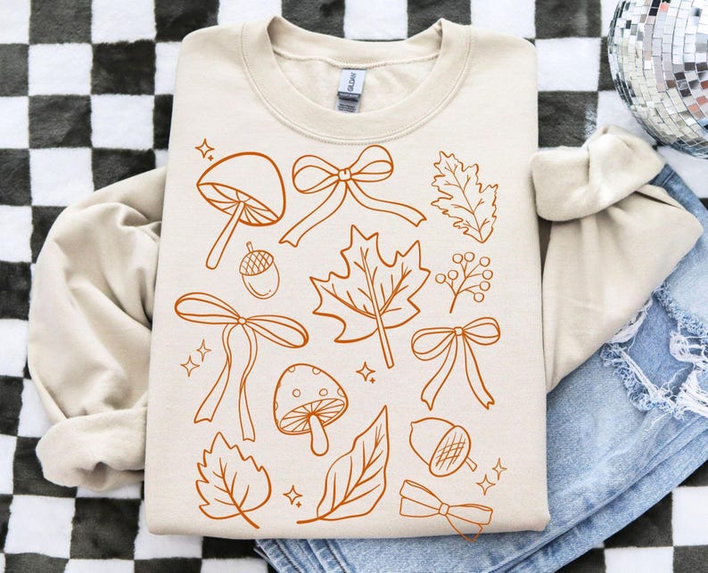Op de afbeelding: Cr&egrave;mekleurige sweatshirt met een herhalend patroon van herfstthema-illustraties in een warme bruine tint. Het ontwerp bevat paddenstoelen, strikken, bladeren, eikels en bessen. De sweatshirt ligt op een spijkerbroek.