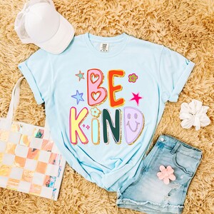 May include: A light blue t-shirt with the words "BE KIND" in colorful, playful lettering. The design includes stars, hearts, and a smiley face. The shirt is paired with denim shorts, a white cap, and a tote bag.