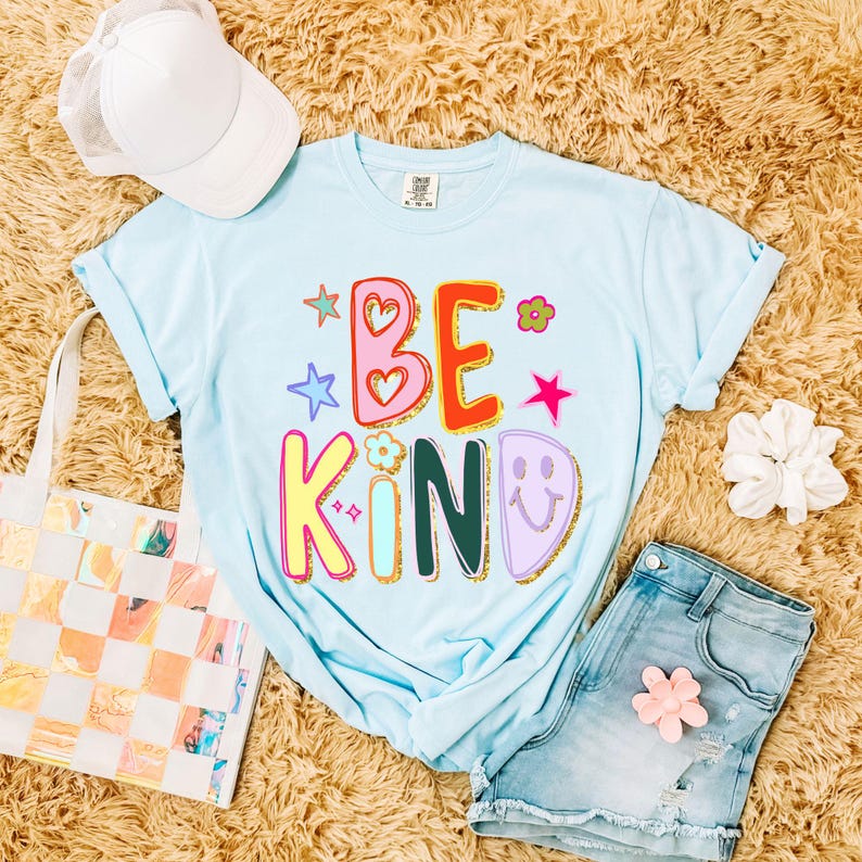 May include: A light blue t-shirt with the words "BE KIND" in colorful, playful lettering. The design includes stars, hearts, and a smiley face. The shirt is paired with denim shorts, a white cap, and a tote bag.