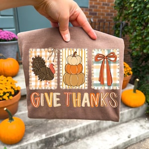 May include: A brown t-shirt featuring three fall-themed designs: a turkey, stacked pumpkins, and a bow. The words "GIVE THANKS" are printed in orange. The shirt is held up by a hand.
