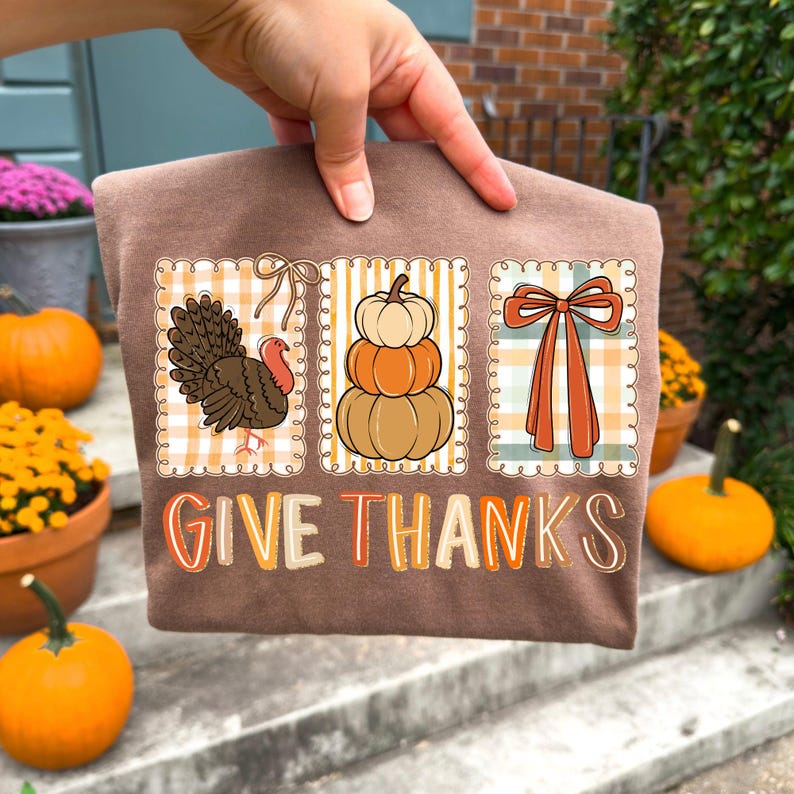 May include: A brown t-shirt featuring three fall-themed designs: a turkey, stacked pumpkins, and a bow. The words "GIVE THANKS" are printed in orange. The shirt is held up by a hand.