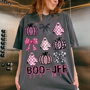Może przedstawiać: Ciemnoszara koszulka z motywem Halloween. Projekt zawiera r&oacute;żowe dynie w panterkę, kokardy i duszki. Tekst "BOO-JEE" jest wydrukowany na czarno z r&oacute;żowymi, ociekającymi akcentami.