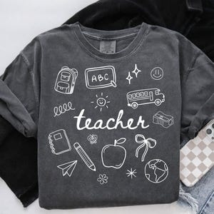 May include: A dark grey t-shirt with a white graphic design of school-themed doodles, including a backpack, an apple, a pencil, a bus, and the word "teacher".