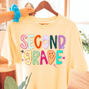Puede incluir: Una camiseta amarillo claro con las palabras "SECOND GRADE" en letras coloridas y divertidas. El dise&ntilde;o incluye estrellas, corazones y una carita sonriente. La camiseta est&aacute; colgada en una percha de madera.