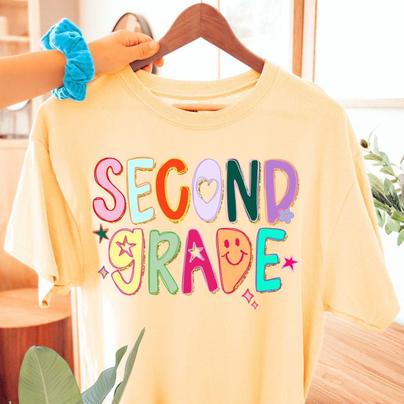 Puede incluir: Una camiseta amarillo claro con las palabras "SECOND GRADE" en letras coloridas y divertidas. El dise&ntilde;o incluye estrellas, corazones y una carita sonriente. La camiseta est&aacute; colgada en una percha de madera.