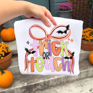 May include: White sweatshirt with a colorful Halloween-themed design. The design features the words "trick or teach" in orange, green, and purple, with bat and bow graphics. The sweatshirt is held up by a hand.