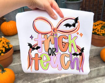 Puede incluir: Sudadera blanca con un diseño colorido con temática de Halloween. El diseño presenta las palabras "trick or teach" en naranja, verde y morado, con murciélagos y lazos. La sudadera es sostenida por una mano.