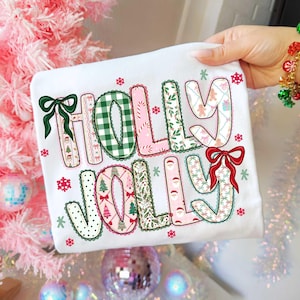 May include: White t-shirt with the words "HOLLY JOLLY" in a festive design. The letters are decorated with Christmas patterns, bows, and snowflakes. The shirt is held by a hand with a colorful bracelet.