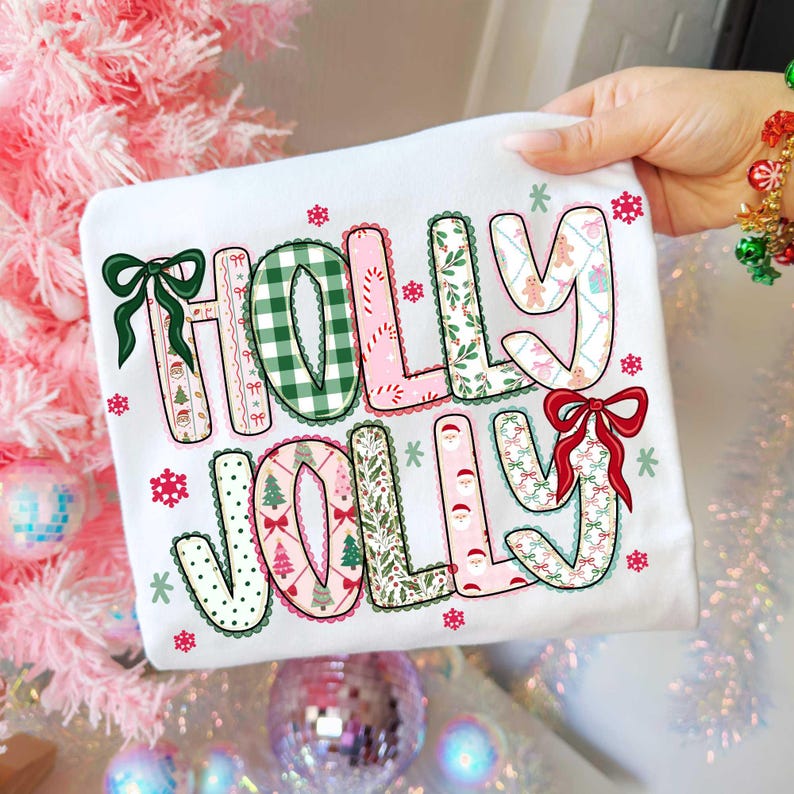 May include: White t-shirt with the words "HOLLY JOLLY" in a festive design. The letters are decorated with Christmas patterns, bows, and snowflakes. The shirt is held by a hand with a colorful bracelet.