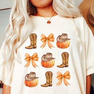 Op de afbeelding: Cr&egrave;mekleurig T-shirt met een aquarelontwerp van cowboylaarzen, pompoenen met cowboyhoeden en oranje strikken. Het ontwerp wordt herhaald over de voorkant van het shirt, waardoor een herfstthema ontstaat.