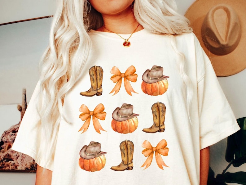 Op de afbeelding: Cr&egrave;mekleurig T-shirt met een aquarelontwerp van cowboylaarzen, pompoenen met cowboyhoeden en oranje strikken. Het ontwerp wordt herhaald over de voorkant van het shirt, waardoor een herfstthema ontstaat.