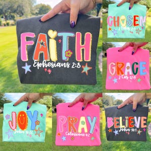 May include: A collection of colorful graphic t-shirts. The shirts feature words like "FAITH", "CHOSEN", "GRACE", "JOY", "PRAY", and "BELIEVE" in vibrant, retro-style lettering. Each shirt includes a Bible verse reference and decorative elements like stars and flowers.