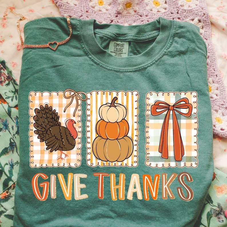 May include: A teal Comfort Colors t-shirt featuring three fall-themed graphics: a turkey, stacked pumpkins, and a bow. The words "GIVE THANKS" are printed in orange and white. A gold necklace with a heart pendant is visible.