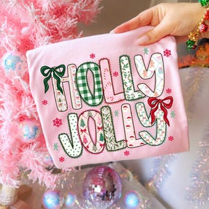 May include: A light pink t-shirt with the words "HOLLY JOLLY" in a festive design. The letters are decorated with patterns, bows, and Christmas-themed illustrations. The shirt is surrounded by snowflakes and Christmas ornaments.