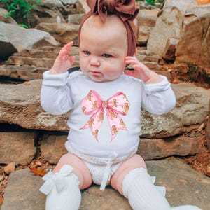 May include: A white long-sleeved baby bodysuit featuring a large pink bow with gingerbread man designs. The baby is wearing a brown headband and white socks with bows. The background is a natural stone setting.