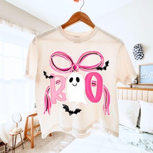 May include: A cream-colored t-shirt with a Halloween-themed design. The design features the word "BOO" in pink letters, a white ghost, a pink bow, and black bats. The shirt is hanging on a wooden hanger.