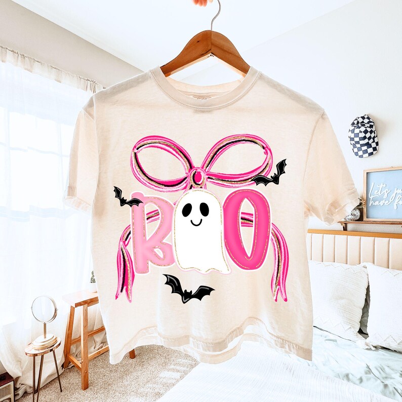 May include: A cream-colored t-shirt with a Halloween-themed design. The design features the word "BOO" in pink letters, a white ghost, a pink bow, and black bats. The shirt is hanging on a wooden hanger.