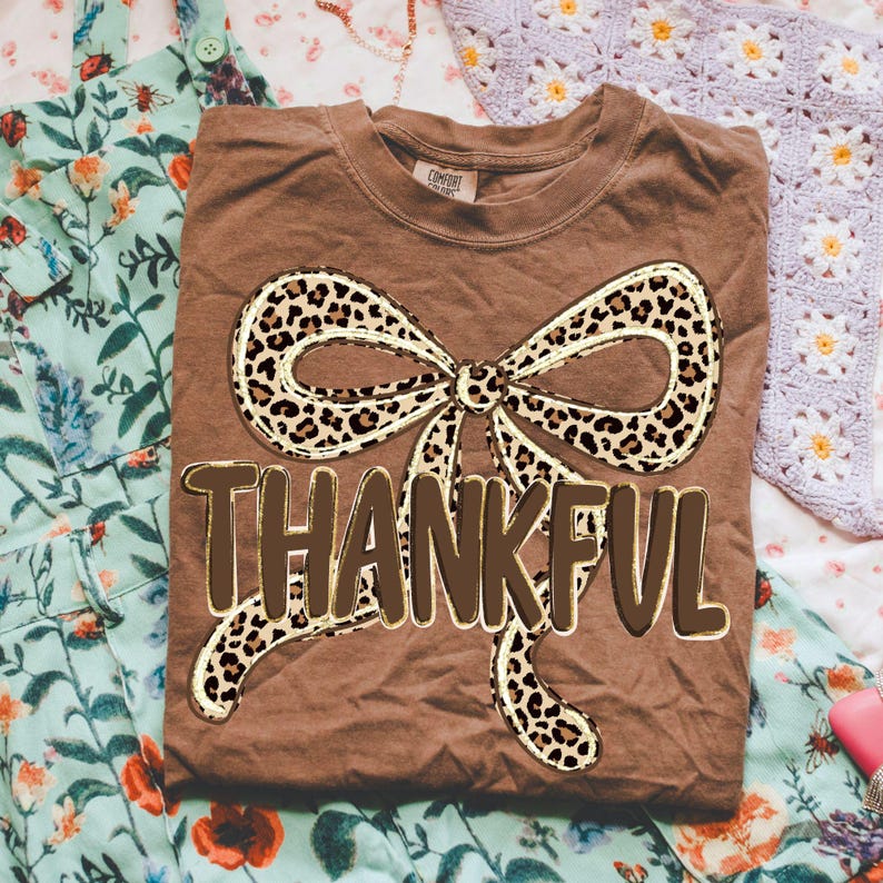 May include: A brown t-shirt with a large bow design featuring a leopard print pattern. The word "THANKFUL" is printed in a brown, bold font below the bow. The shirt is folded and rests on a floral patterned fabric.