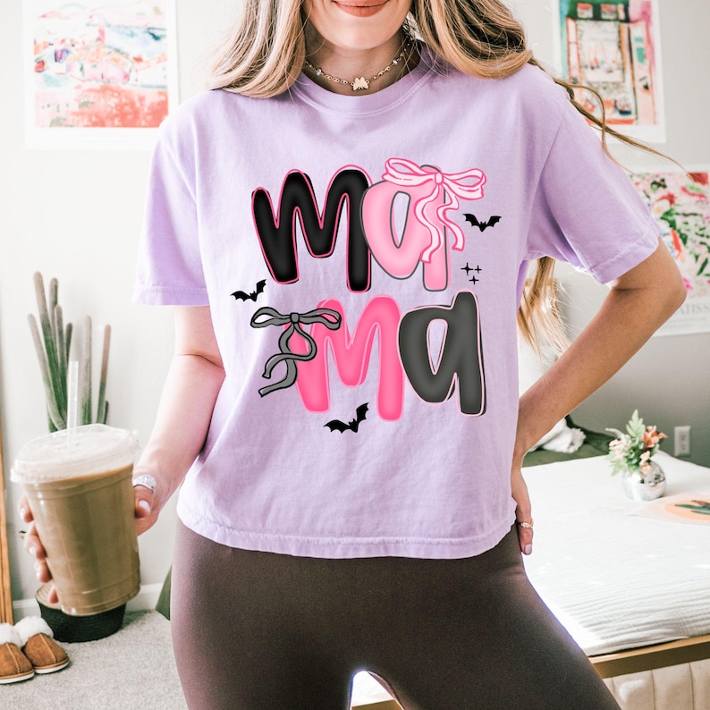 May include: A light purple t-shirt with the word "Mama" in black and pink, decorated with bows and bats. The shirt is a casual style, perfect for everyday wear.