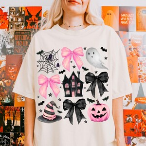 May include: Cream-colored t-shirt featuring Halloween-themed illustrations. The design includes pink bows, a haunted house, a jack-o'-lantern, a witch's hat, bats, stars, a spiderweb, and a ghost. The text "GIRLS JUST WANNA HAVE FUN" is visible on the background.