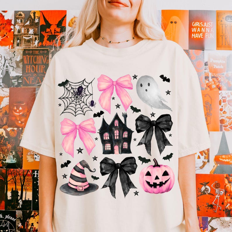 May include: Cream-colored t-shirt featuring Halloween-themed illustrations. The design includes pink bows, a haunted house, a jack-o'-lantern, a witch's hat, bats, stars, a spiderweb, and a ghost. The text "GIRLS JUST WANNA HAVE FUN" is visible on the background.