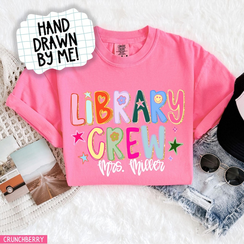 Może przedstawiać: R&oacute;żowa koszulka z napisem "LIBRARY CREW" w kolorowych, fantazyjnych literach. Koszulka zawiera r&oacute;wnież tekst "Mrs. Miller" i frazę "HAND DRAWN BY ME!" w dymku. Koszulka jest złożona na białej powierzchni.