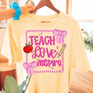 Puede incluir: Camiseta amarillo claro con las palabras "Teach Love Inspire" en rosa, una manzana roja, lazos rosas y un l&aacute;piz. La camiseta est&aacute; colgada en una percha de madera.
