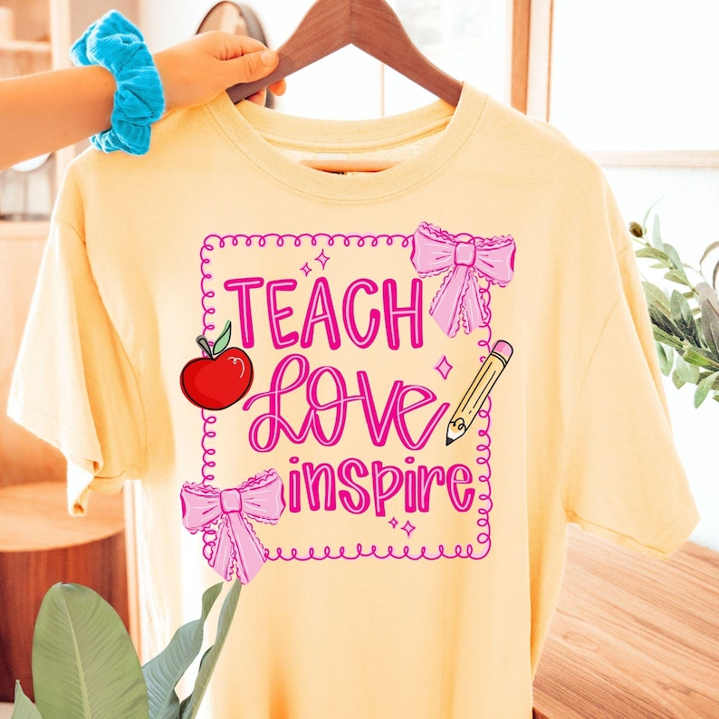 Puede incluir: Camiseta amarillo claro con las palabras "Teach Love Inspire" en rosa, una manzana roja, lazos rosas y un l&aacute;piz. La camiseta est&aacute; colgada en una percha de madera.