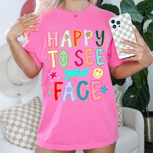 May include: A bright pink t-shirt with the phrase "HAPPY TO SEE YOUR FACE" in colorful, playful lettering. The design includes a flower, stars, and smiley faces. The shirt is a casual, comfortable style.
