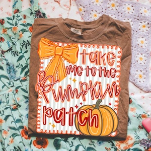 May include: Brown t-shirt with the text "Take Me To The Pumpkin Patch" in red and orange. The design includes an orange bow and a pumpkin illustration. The shirt is folded on a floral patterned surface.